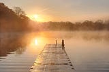 In the middle, a dock extending into the ripples and mists of the water of the lake, the sky is alit by the warm sun on the left rising over the tree line.