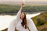 Angel with wavy brunnette hair and white clothing raising a glass of white wine. She is standing on a hill overlooking a river. The background is blurred, but the lighting gives a sense of serence peace to the overall picture.