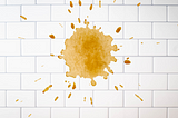 A coffee stain splashed against white subway tiles, creating a warm sepia explosion with scattered droplets radiating outward.