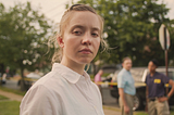 A close-up shot of a woman. We can see from her shoulders up. She is wearing a plain white shirt. Her body is aimed to the right, but her face is turned towards to the camera. She has blonde hair. Behind her, blurred out, are two FBI officers, and behind them a car. The woman is not smiling, her face is solemn and expressionless.