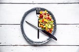 A fork and knife on a round plate in the position of clock hands, with salad in between as a symbol for intermittent fasting.