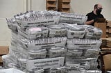 Stacks of mail cartons full of ballots being sorted by a person in the background wearing a face mask.