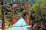 Old rusty single-lane gravel-covered suspension bridge with weight limit limit sign reading maximum of three-tons and an orange warning sign noting maximum height of thirteen feet, eight inches. The image was taken straight on down the center of the roadway with the thick bradded wires projecting out from the bridge to out of sight past the photographer. The bridge road empties onto a single road into a steep hillside thickly covered by trees and brush. The true length distance is deceiving.