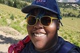 Author on top of a mountain with green hills rolling behind them leading toward a mountain top crested with clouds on a sunny day. Aigner Loren Wilson is smiling wearing yellow sunglasses and a jean NASA hat. Author’s life. Life of a publishing speculative fiction author. Speculative Fiction author. Aigner Loren Wilson