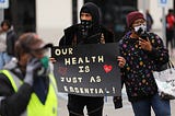 A photo of Amazon workers protesting. One is holding up a sign that says “Our health is just as essential.”