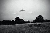 Grainy black-and-white photo of a UFO flying over a park bench overlooking an urban scene in the distance.
