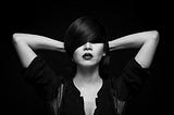 A black and white image of a woman with her eyes covered by her hair. She holds her hands behind her head, partly obscuring her face, conveying an air of introspection and mystery. The dark background emphasizes her expression and the moody, somber tone of the photograph, resonating with themes of complexity, inner conflict, and profound emotion.