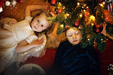 Two young children, seen from above, lie on a parquet floor beneath a decorated Christmas tree with warm lights. One gazes into the camera, the other has their eyes closed. Sparkly snowfall overlays the scene.
