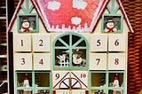 A wooden advent calendar in the shape of a house. There are little windows with snowpeople peeking out, and there is glittering snow painted on the roof.