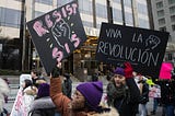 Marchers hold signs that say “Resist Sis” and “Viva La Revolucion” in front of Trump International Hotel on January 19, 2019