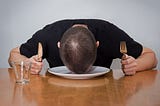 A person holding a fork and knife at a dinner table, resting their head on an empty plate.