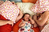 Exhausted parents covering their faces with pillows as baby cries between them.