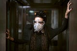 A woman wearing a dark costume and a mask is posing inside an elevator.