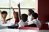 A photo of a young black boy with his hand raised at his desk.