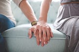 Closeup of two people sitting next to each other on a pale blue sofa in a brightly-lit room. One’s hand rests on the other’s hand, which rests palm-down on the cushion between them.