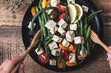 Hands using wooden utensils to toss a bowl of green beans, cherry tomatoes, and grilled tofu cubes in sauce/vinaigrette.