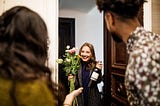 A woman holding a bouquet and a wine bottle in their neighbor’s doorway.