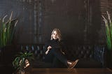A woman wearing black is sitting on a black couch while holding a tablet.