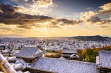 Sunset view over Matsuyama City from the rooftops of Matsuyama Castle, with golden light spilling across the skyline and the Seto Inland Sea glowing in the distance.