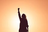 Woman as shadow figure raising the fist in the air and an orange sunny background