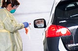 A photo of a man sitting in his car wearing a mask waiting to get tested by a health care worker.