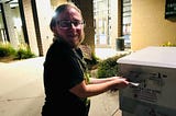 The author is putting his ballot in a drop box. He is wearing a black t-shirt and has dark-rimmed glasses. It’s nighttime. He looks anxious and excited.