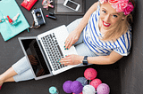 Bird’s Eye view of a blond woman looking up. She is sitting on the ground with a silver laptop on her lap surrounded by a teal folder, a camera, iPad, cell phone, and piles of colorful balls of yarn. From Find Your Niche On Medium by Gema Mora and Canopy Invocations