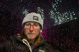 The author is surrounded by white and purple lights. He’s wearing a knitted hat that says “Joy as resistance.” He’s wearing black rimmed glasses and has a beard with enough white to betray that he’s middle-aged. He has long brown hair and a black coat that says “Denver” over a purple sweater. Purple glitter eyeliner matches the lights behind him.