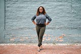A photo of a confident black woman standing against a blue brick background in a power pose.