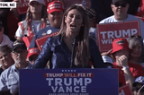 Alina Habba yelling into a microphone at a rally behind a Trump/Vance sign