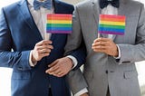 Gay male couple in wedding suites with arms interlocked hold little gay pride flags.
