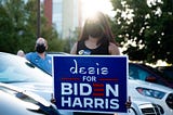 A woman wearing face mask holds a sign that says “desis for Biden Harris.”
