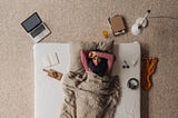 An aerial photo of a sick woman lying on a mattress on the floor. A laptop, books, tissues, etc. are strewn on ground.