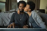 A photo of a black couple. He is playing keyboard and she kisses him on the cheek.