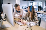 An employee working at her desk with headphones in is interrupted by a manager who comes by for a chat.