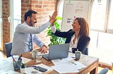 A businesswoman and businessman are high-fiving across a table from each other, both with smiles on their faces