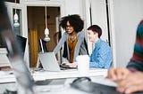 Two women entrepreneurs work together. One woman sits at her desk and the other is teaches  her something off her laptop.