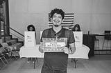 The image shows a man smiling while holding a sign that reads, “Your Vote Matters 2024.” He is standing in what appears to be a polling station, with other people in the background casting their votes in booths. An American flag is visible in the background, emphasizing the patriotic setting. The image conveys a positive and encouraging message about the importance of voting in the 2024 election