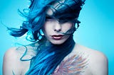 A non-binary person with long, blue hair and black contact lenses stares intensely into the camera lens. They have a colorful tattoo of wings on their chest, and the background is light blue.