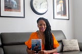 Martinique Lewis sitting on a black couch holding her book on her lap.