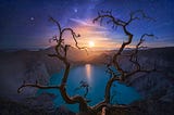 A fine art photograph of bare tree branches framing a moon and lake setting with purple clouds and stars
