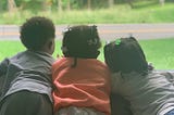 3 young Black children seen from behind, looking out the window at grass and a road.