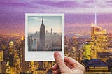 A hand holding a Polaroid photo of NYC up to the NYC skyline at evening.