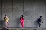 3 elementary school students wearing face masks and standing against a cement wall, 6 feet of distance between them.