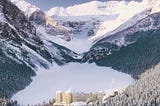 A hotel in a snowy valley with snow-covered evergreen trees in the foreground and snowy mountains in the background.