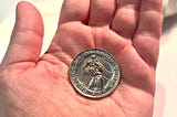 Color vertical closeup image, against a white porcelain countertop background, of the caucausian author’s left reddish-looking skin hand’s palm facing up holding within its center a silver coin stating “PROTECT US ST. CHRISTOPHER”, showing a traditional medallion of the patron Saint carrying a child on his back, three stars at the bottom, and strobes of light engraved coming from all around him.