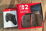 A Switch 2 Pro Controller Box (left) and a Switch 2 Box (right) sitting next to each other on my slightly ugly patterned couch.