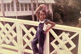 A young girl in blue overalls leans against a white fence. She is doing a ballet pose with one arm curved over her head and the other on her hip. One leg is bent. She’s got a big open-mouth smile.