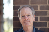 Outdoor portrait of William Moyers standing by a brick pillar. He is wearing a blue checked blazer over a denim button-down shirt.