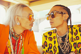 The image depicts two men engaged in an intense conversation. Both are dressed in vibrant, eclectic clothing with bold colors and patterns. The man on the left has long, white hair and wears an orange outfit adorned with various necklaces and beads. He also wears orange-tinted sunglasses and has large, decorative earrings. The man on the right has braided hair and wears a yellow jacket with a leopard print design. He is accessorized with multiple chains around his neck.